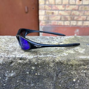Op de afbeelding: Zwarte zonnebril met blauwe glazen op een betonnen oppervlak. De zonnebril heeft een strak, modern design met een gebogen montuur en een klein zilveren embleem op de pootjes. De glazen zijn helderblauw.
