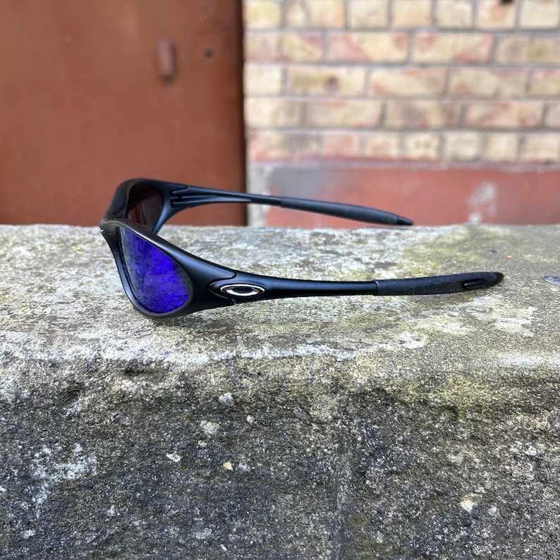 Op de afbeelding: Zwarte zonnebril met blauwe glazen op een betonnen oppervlak. De zonnebril heeft een strak, modern design met een gebogen montuur en een klein zilveren embleem op de pootjes. De glazen zijn helderblauw.