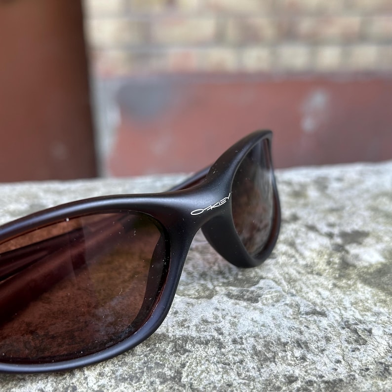 May include: Close-up of black Oakley sunglasses with brown tinted lenses. The Oakley logo is visible on the temple. The sunglasses have a curved, sporty design.
