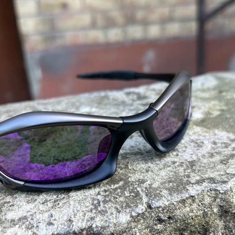 May include: Close-up of a pair of sunglasses with a black frame and purple lenses. The sunglasses have a sleek, modern design. The lenses reflect the surrounding environment.