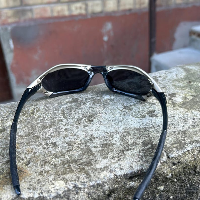 May include: Sunglasses with a silver frame and black lenses. The sunglasses have a sleek, modern design with a curved shape. The arms are black and extend back from the frame. The sunglasses are resting on a rough, gray surface.
