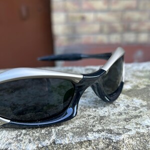 May include: Close-up of black sunglasses with silver frames. The sunglasses have dark lenses and a sleek, modern design. The frames are a combination of black and silver, creating a stylish contrast. The sunglasses are resting on a textured surface.