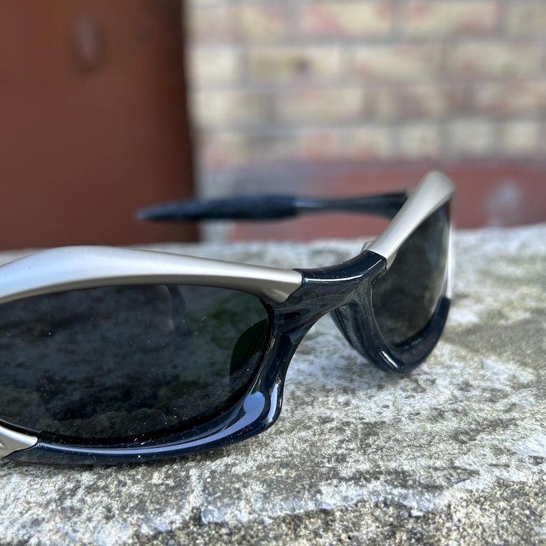 May include: Close-up of black sunglasses with silver frames. The sunglasses have dark lenses and a sleek, modern design. The frames are a combination of black and silver, creating a stylish contrast. The sunglasses are resting on a textured surface.