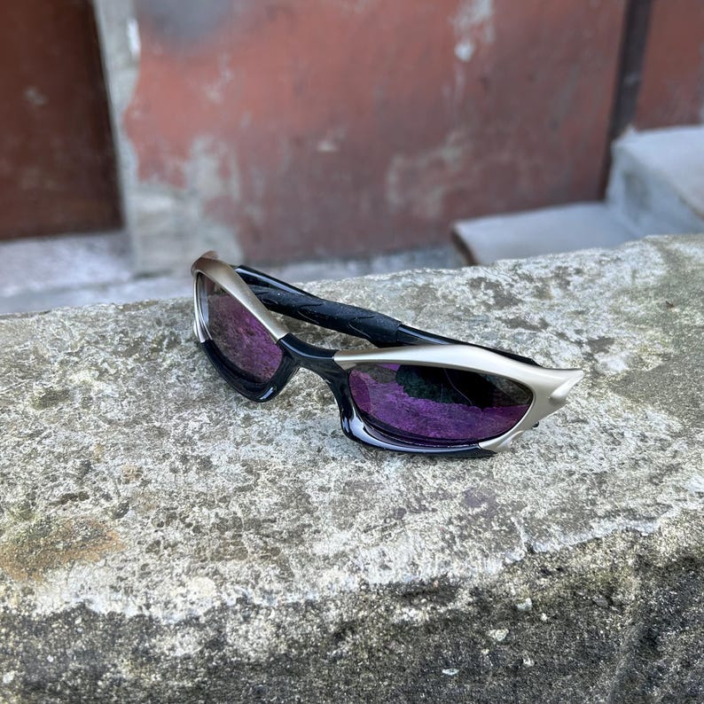 Puede incluir: Gafas de sol elegantes con montura negra y detalles plateados. Las lentes tienen un tinte p&uacute;rpura. Las gafas de sol est&aacute;n sobre una superficie gris texturizada, con un fondo borroso rojo y gris.