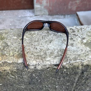 May include: Sunglasses featuring a black frame and brown lenses. The arms have a brown, leather-effect finish. The glasses are positioned on a weathered concrete surface. The design is sporty and contemporary.