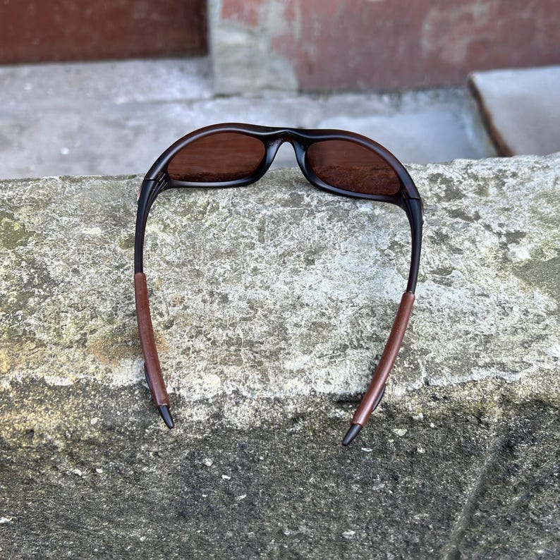 May include: Sunglasses featuring a black frame and brown lenses. The arms have a brown, leather-effect finish. The glasses are positioned on a weathered concrete surface. The design is sporty and contemporary.