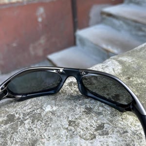 May include: Black sunglasses with a sleek, modern design. The sunglasses have dark lenses and a glossy black frame. The arms of the sunglasses curve slightly for a comfortable fit. The sunglasses are resting on a stone surface.