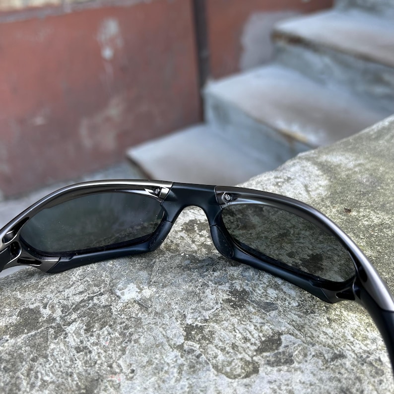 May include: Black sunglasses with a sleek, modern design. The sunglasses have dark lenses and a glossy black frame. The arms of the sunglasses curve slightly for a comfortable fit. The sunglasses are resting on a stone surface.