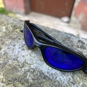 Op de afbeelding: Zwarte zonnebril met blauwe glazen. De zonnebril heeft een strak ontwerp, met de merknaam "Oakley" op het montuur. De glazen reflecteren een levendige blauwe kleur, die contrasteert met het zwarte montuur.