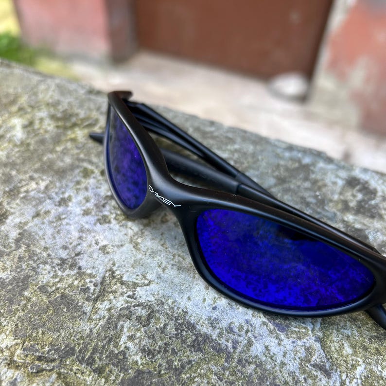 Op de afbeelding: Zwarte zonnebril met blauwe glazen. De zonnebril heeft een strak ontwerp, met de merknaam "Oakley" op het montuur. De glazen reflecteren een levendige blauwe kleur, die contrasteert met het zwarte montuur.