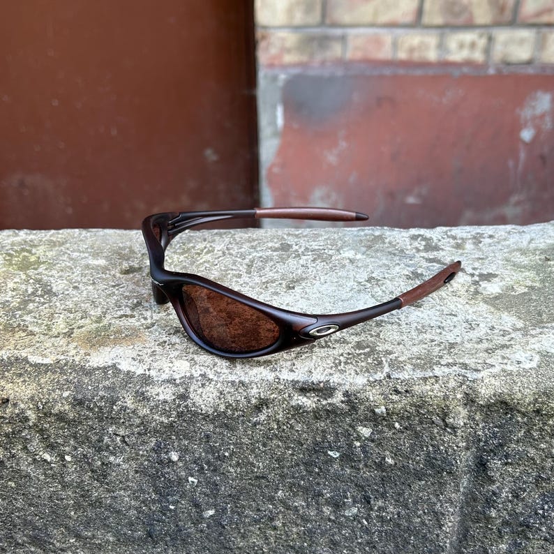 May include: Brown sunglasses with a curved frame and brown lenses. The arms of the sunglasses have a dark brown colour with a lighter brown tip. The sunglasses are resting on a grey, textured surface.