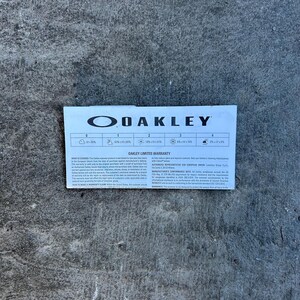 Op de afbeelding: Een witte en blauwe Oakley Limited Warranty kaart. De kaart toont het Oakley-logo en tekst met garantie-informatie. De kaart ligt op een grijs betonnen oppervlak.