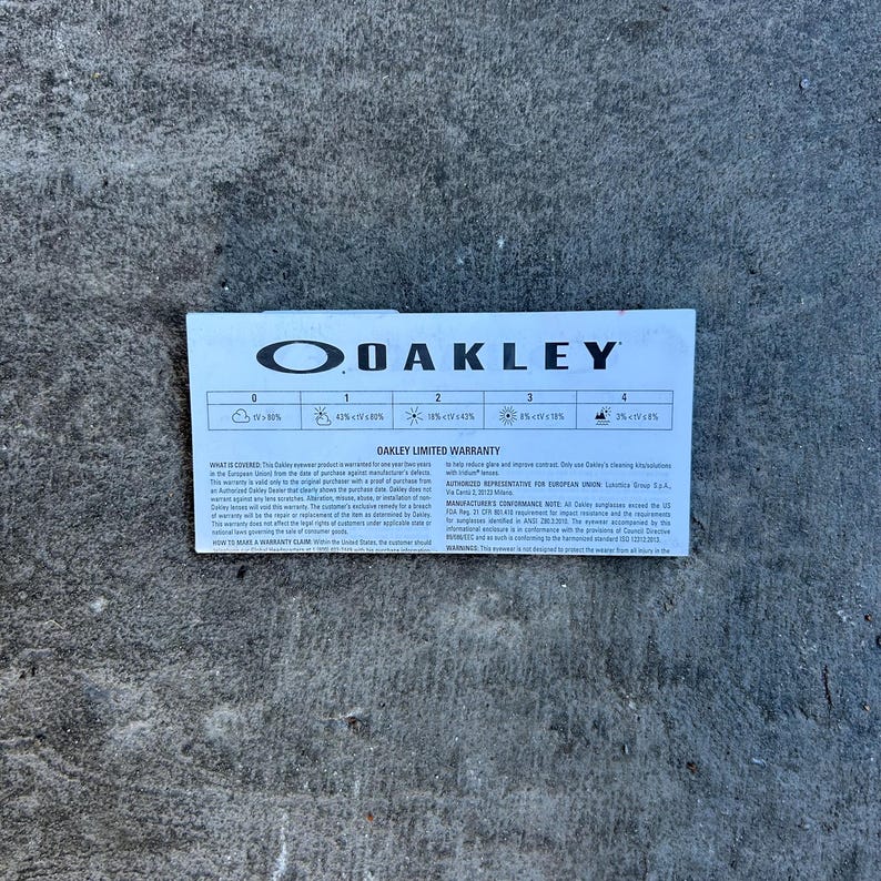 Op de afbeelding: Een witte en blauwe Oakley Limited Warranty kaart. De kaart toont het Oakley-logo en tekst met garantie-informatie. De kaart ligt op een grijs betonnen oppervlak.