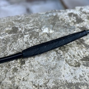 Puede incluir: Primer plano de un objeto negro con el texto "MADE IN U.S.A." en relieve. El objeto parece estar hecho de un material flexible y est&aacute; colocado sobre una superficie de piedra gris texturizada. El objeto es largo y delgado.
