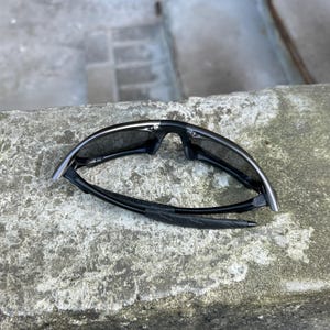 May include: Black sport sunglasses with a curved frame and dark lenses. The sunglasses are folded and rest on a textured, gray surface. The arms of the sunglasses have a padded section for comfort.