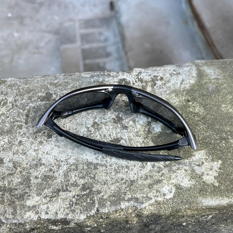 May include: Black sport sunglasses with a curved frame and dark lenses. The sunglasses are folded and rest on a textured, gray surface. The arms of the sunglasses have a padded section for comfort.
