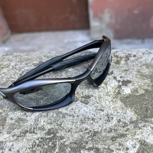 May include: Black and gray sunglasses with a sleek, modern design. The sunglasses feature a curved frame and dark lenses. The temples are black and textured, providing a secure fit. The overall aesthetic is sporty and stylish.