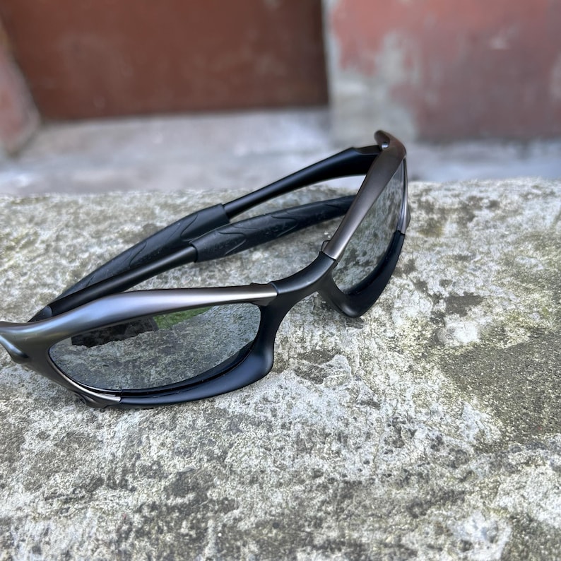 May include: Black and gray sunglasses with a sleek, modern design. The sunglasses feature a curved frame and dark lenses. The temples are black and textured, providing a secure fit. The overall aesthetic is sporty and stylish.
