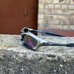 Puede incluir: Gafas de sol plateadas y negras con lentes tintadas de p&uacute;rpura. Las gafas de sol tienen un dise&ntilde;o elegante y moderno con una montura curvada y patillas negras. Las lentes reflejan un tono p&uacute;rpura.