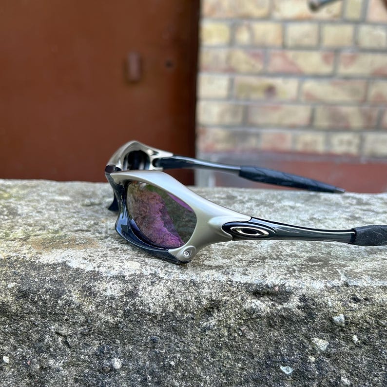 Puede incluir: Gafas de sol plateadas y negras con lentes tintadas de p&uacute;rpura. Las gafas de sol tienen un dise&ntilde;o elegante y moderno con una montura curvada y patillas negras. Las lentes reflejan un tono p&uacute;rpura.