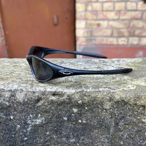 May include: Black sunglasses with a modern design. The sunglasses have dark lenses and a matte black frame with a silver accent near the hinges. The arms of the sunglasses are thin and curve around the ears.