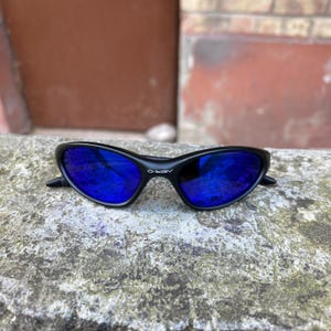 Op de afbeelding: Zwarte zonnebril met blauwe reflecterende glazen. De zonnebril heeft een gebogen montuur en de merknaam "Oakley" is zichtbaar op de rechter poot. De zonnebril ligt op een grijs, getextureerd oppervlak.