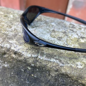 Op de afbeelding: Zwarte zonnebril met een strak, modern design. De zonnebril heeft een gebogen montuur en donkere glazen. Het Oakley-logo is zichtbaar op de pootjes. De zonnebril ligt op een gestructureerd, grijs oppervlak.