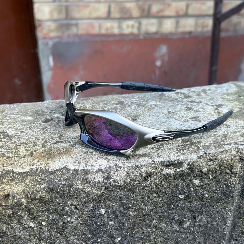 Puede incluir: Gafas de sol plateadas y negras con lentes moradas. Las gafas tienen un dise&ntilde;o elegante y moderno con una montura curvada y patillas negras. Las lentes son de color morado vibrante, ofreciendo un aspecto deportivo y elegante.