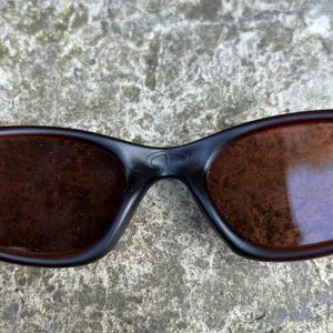 May include: Black sunglasses with brown tinted lenses. The sunglasses have a curved frame and a small logo on the bridge. The lenses are a warm brown colour, and the frame is a matte black.
