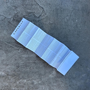 Op de afbeelding: Gevouwen wit papier met het OAKLEY-logo bovenaan. Het papier staat vol met kleine tekst en ligt op een grijze betonnen ondergrond. Het papier is harmonica-achtig gevouwen.