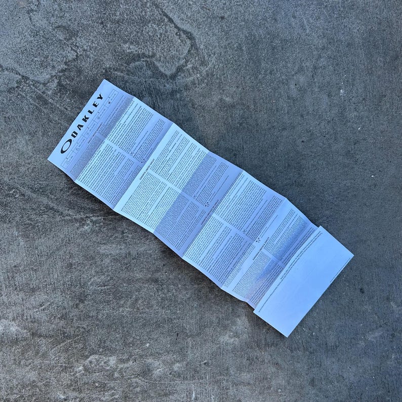 Op de afbeelding: Gevouwen wit papier met het OAKLEY-logo bovenaan. Het papier staat vol met kleine tekst en ligt op een grijze betonnen ondergrond. Het papier is harmonica-achtig gevouwen.