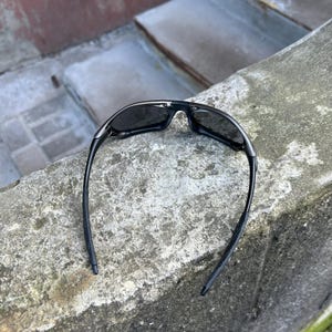 May include: Black sunglasses with a sleek, modern design. The sunglasses have dark lenses and a black frame. The arms of the sunglasses are thin and curve down. The sunglasses are resting on a concrete surface.