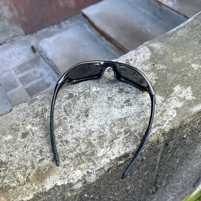 May include: Black sunglasses with a sleek, modern design. The sunglasses have dark lenses and a black frame. The arms of the sunglasses are thin and curve down. The sunglasses are resting on a concrete surface.