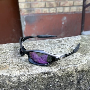 May include: Black sunglasses with purple lenses rest on a concrete surface. The sunglasses have a sleek, curved design with dark frames and textured arms. The lenses reflect a vibrant purple hue.