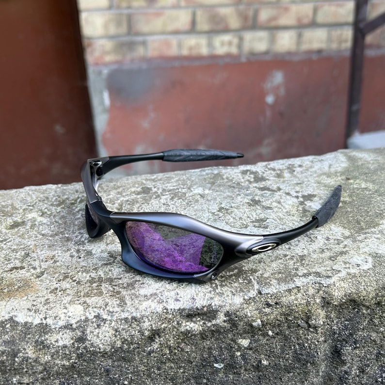 May include: Black sunglasses with purple lenses rest on a concrete surface. The sunglasses have a sleek, curved design with dark frames and textured arms. The lenses reflect a vibrant purple hue.