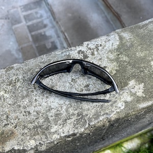 May include: Black sunglasses with a sleek, modern design. The sunglasses have dark lenses and a black frame with silver accents. The arms are thin and flexible. The sunglasses are resting on a rough, gray surface.