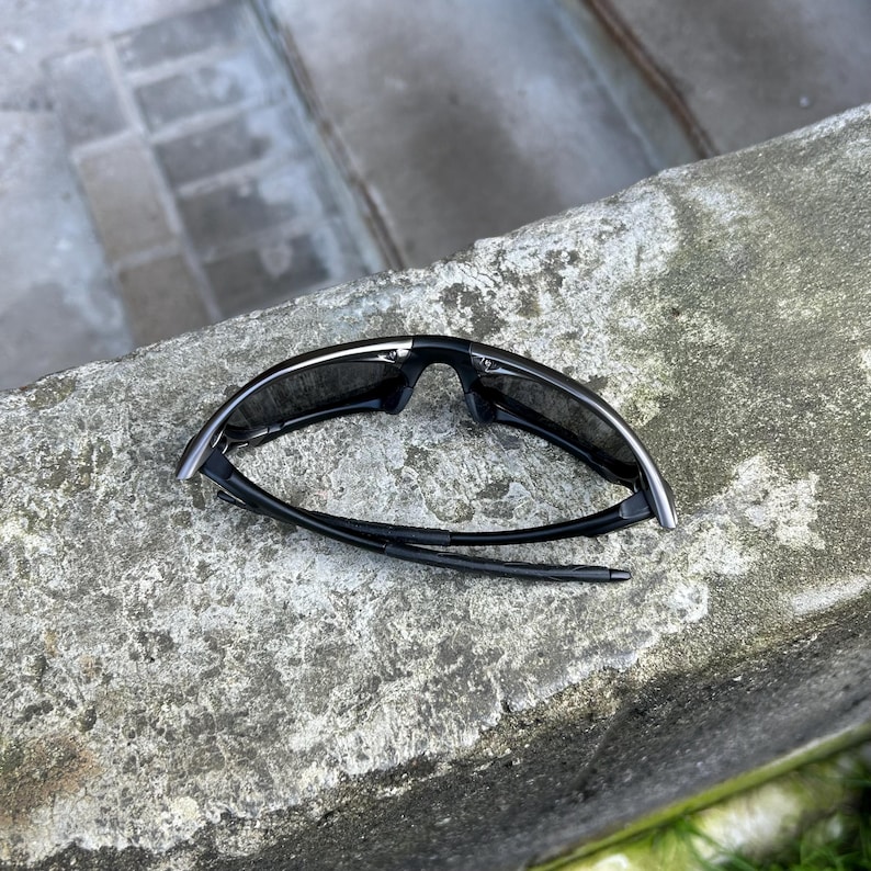 May include: Black sunglasses with a sleek, modern design. The sunglasses have dark lenses and a black frame with silver accents. The arms are thin and flexible. The sunglasses are resting on a rough, gray surface.