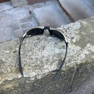 May include: Black sunglasses with a silver frame and dark lenses. The sunglasses have a sleek, modern design with curved temples. The image shows the sunglasses resting on a textured, gray surface.