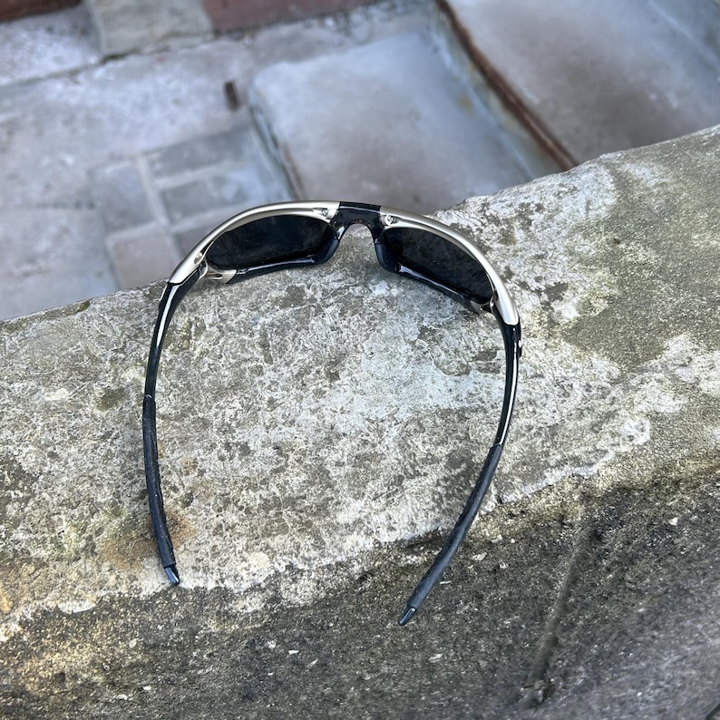 May include: Black sunglasses with a silver frame and dark lenses. The sunglasses have a sleek, modern design with curved temples. The image shows the sunglasses resting on a textured, gray surface.