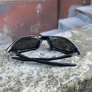 May include: Black sunglasses with a sleek, modern design. The sunglasses have dark lenses and a black frame with silver accents. The arms are folded, revealing the inner details. The sunglasses are resting on a concrete surface.