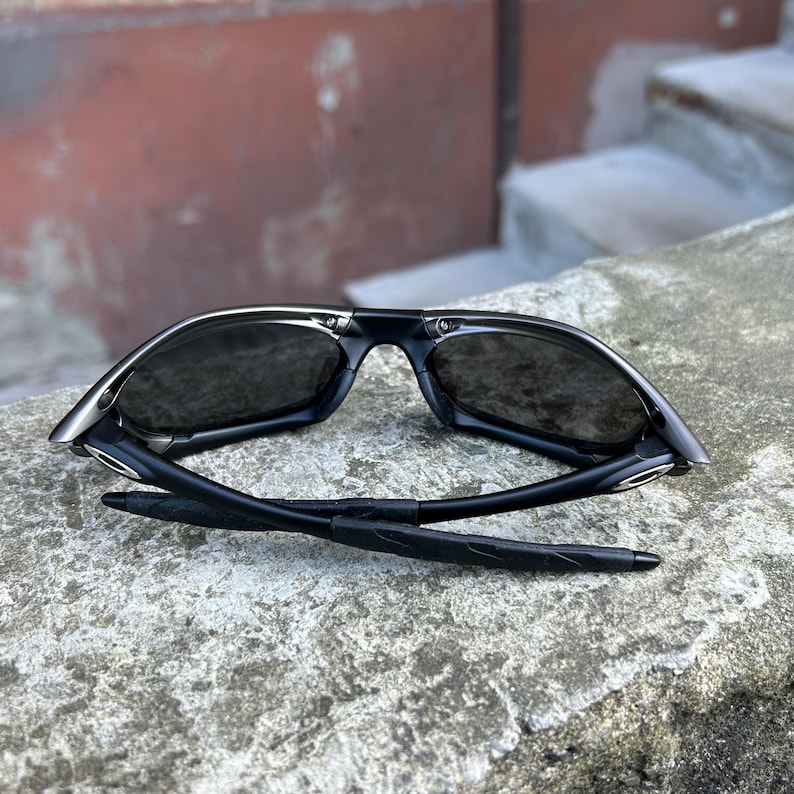 May include: Black sunglasses with a sleek, modern design. The sunglasses have dark lenses and a black frame with silver accents. The arms are folded, revealing the inner details. The sunglasses are resting on a concrete surface.