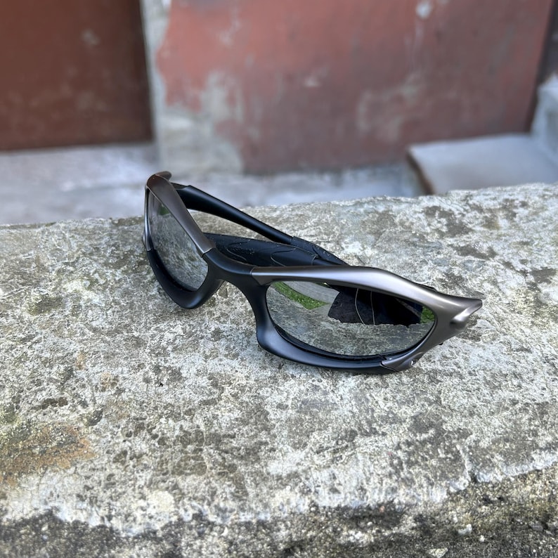 May include: Black and silver sunglasses with a unique, angular design. The sunglasses have a sleek, modern look with dark lenses and a curved frame. The arms are black, and the frame has a silver accent.