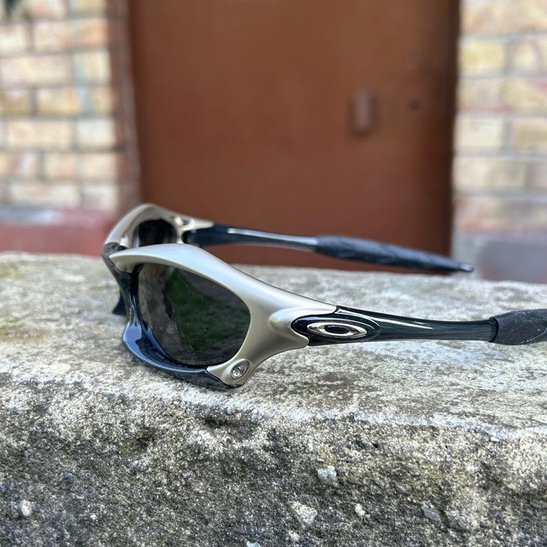 May include: Silver and black sunglasses with dark lenses. The sunglasses have a sleek, modern design with a silver frame and black temples. The lenses are dark, providing shade. The sunglasses are resting on a stone surface.