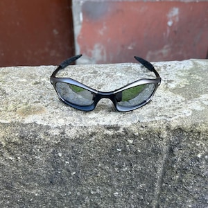May include: Black and silver sunglasses with a curved frame and reflective lenses. The sunglasses are resting on a gray stone surface, with a blurred red and brown background.