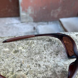 May include: Brown sunglasses with a curved frame and tinted lenses. The temple arms are a darker brown with a leather-like texture. The text "MADE IN U.S.A." is visible on the temple arm.