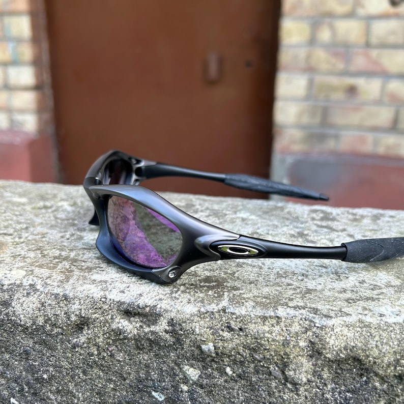 May include: Black sunglasses with purple lenses rest on a gray stone surface. The sunglasses have a sleek, modern design with a matte finish and curved arms. The lenses reflect a purple hue, and the frame features a small silver logo.