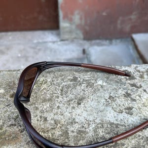 May include: Dark brown sunglasses with a sleek, modern design. The frames are a deep, rich brown, complemented by brown temples. The lenses have a subtle tint, offering a stylish and functional accessory for eye protection.