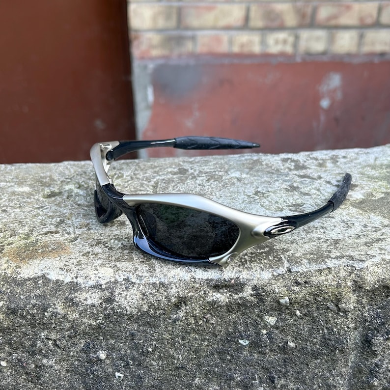 May include: Sleek, black and silver sunglasses with dark lenses. The sunglasses have a curved design with a silver frame and black arms. The Oakley logo is visible on the side of the frame. The sunglasses are resting on a gray, textured surface.