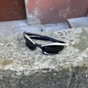 May include: Sleek, modern sunglasses with a silver frame and black lenses. The sunglasses have a unique, angular design. The temples are black. The sunglasses are resting on a gray stone surface.
