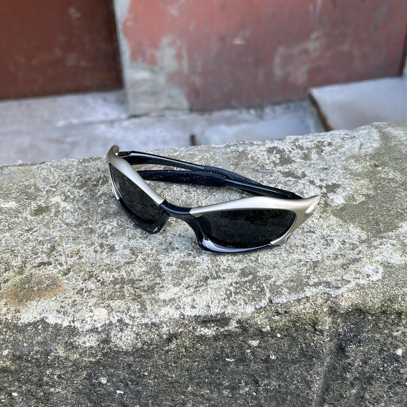 May include: Sleek, modern sunglasses with a silver frame and black lenses. The sunglasses have a unique, angular design. The temples are black. The sunglasses are resting on a gray stone surface.